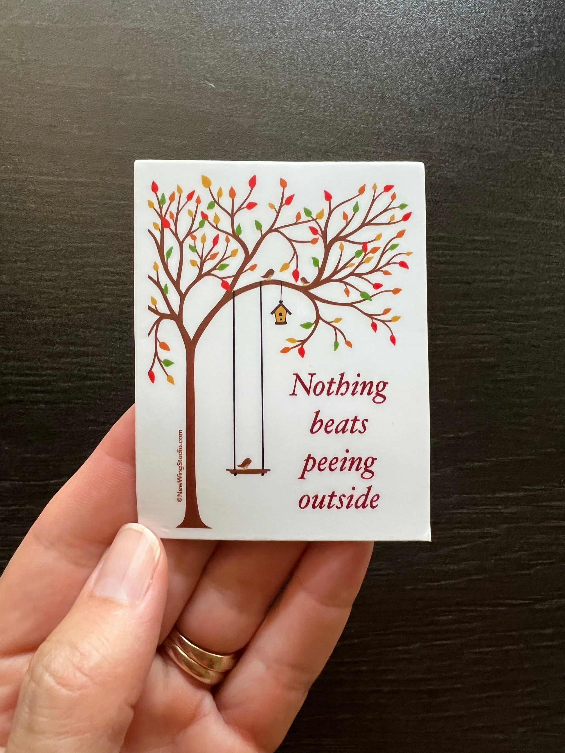 Our "Nothing Beats Peeing Outside" sticker is perfect for those who love the outdoors and have a great sense of humor.  Sure to get a reaction, this whimsical sticker features a serene setting of a tree in fall with a bird on a swing and the contrasting script, "Nothing Beasts Peeing Outside." Perfect for those with a sassy, off-color, irreverent, coarse sense of humor.  This is a picture of the sticker in a human hand.  When nature calls… newwingstudio.com