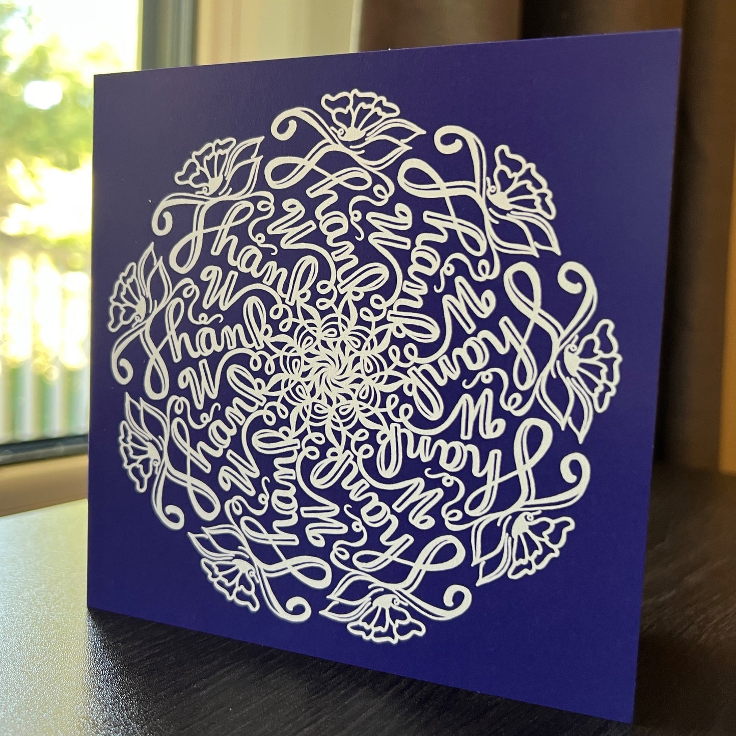 This hidden word Mandala greeting card allows the sender to say “Thank you” in a beautiful and creative Way.  The card has a deep purple background, on which a striking white mandala is printed.  The Mandala features the phrase “Thank U” nine times in a mesmerizing swirling pattern, and the pattern is adorned with flowers, leaves, and vines. This is a picture on a desk with a natural background Newwingstudio.com