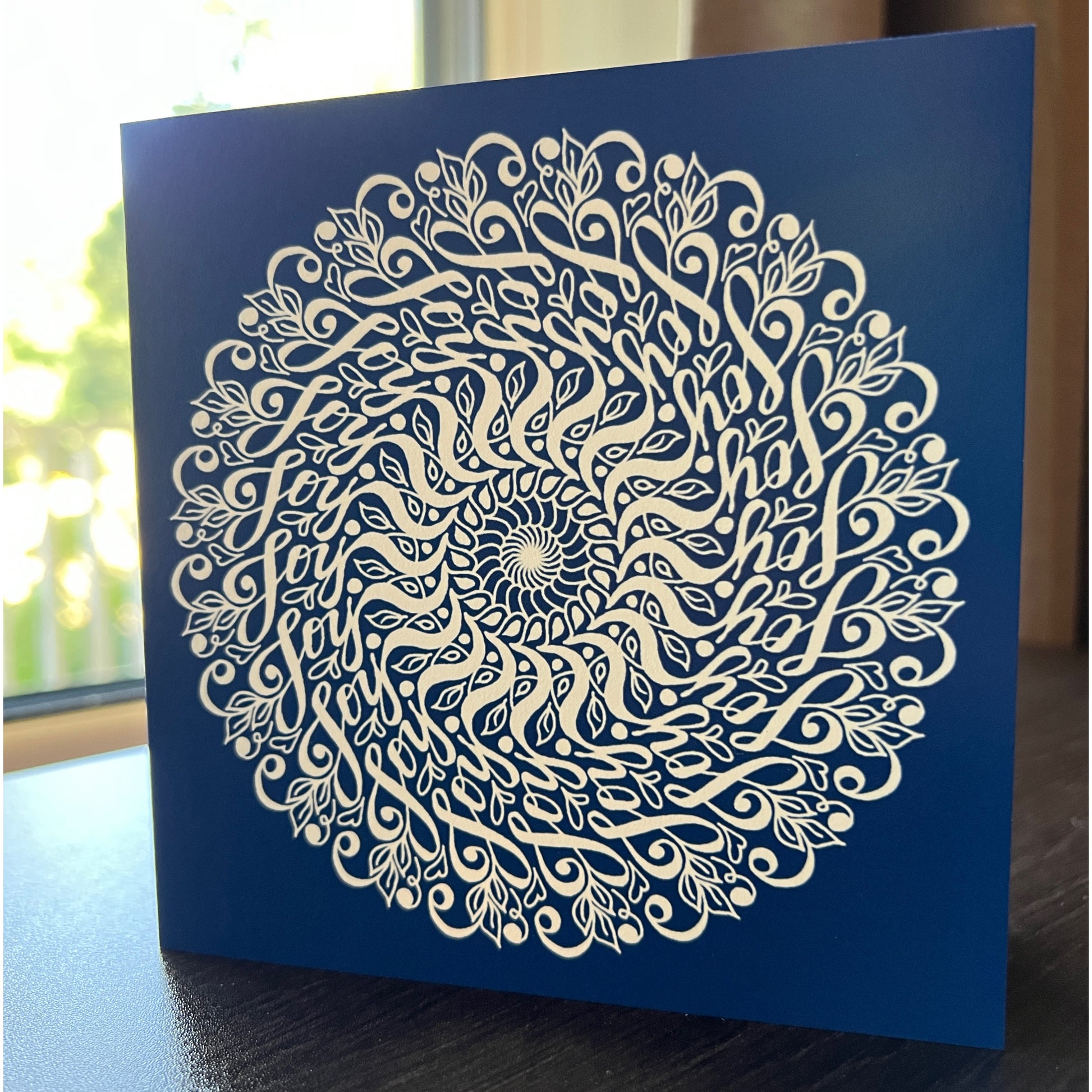 Our "Joy Mandala" greeting card is one of our best sellers and features a deep blue background and a swirling mandala with the hidden word "Joy" incorporated into the design.  You can almost get lost in the movement of this elegant pattern!  This hidden word mandala greeting card is on a desk with a natural background.   Another way to describe it is a hidden message greeting card.  Newwingstudio.com