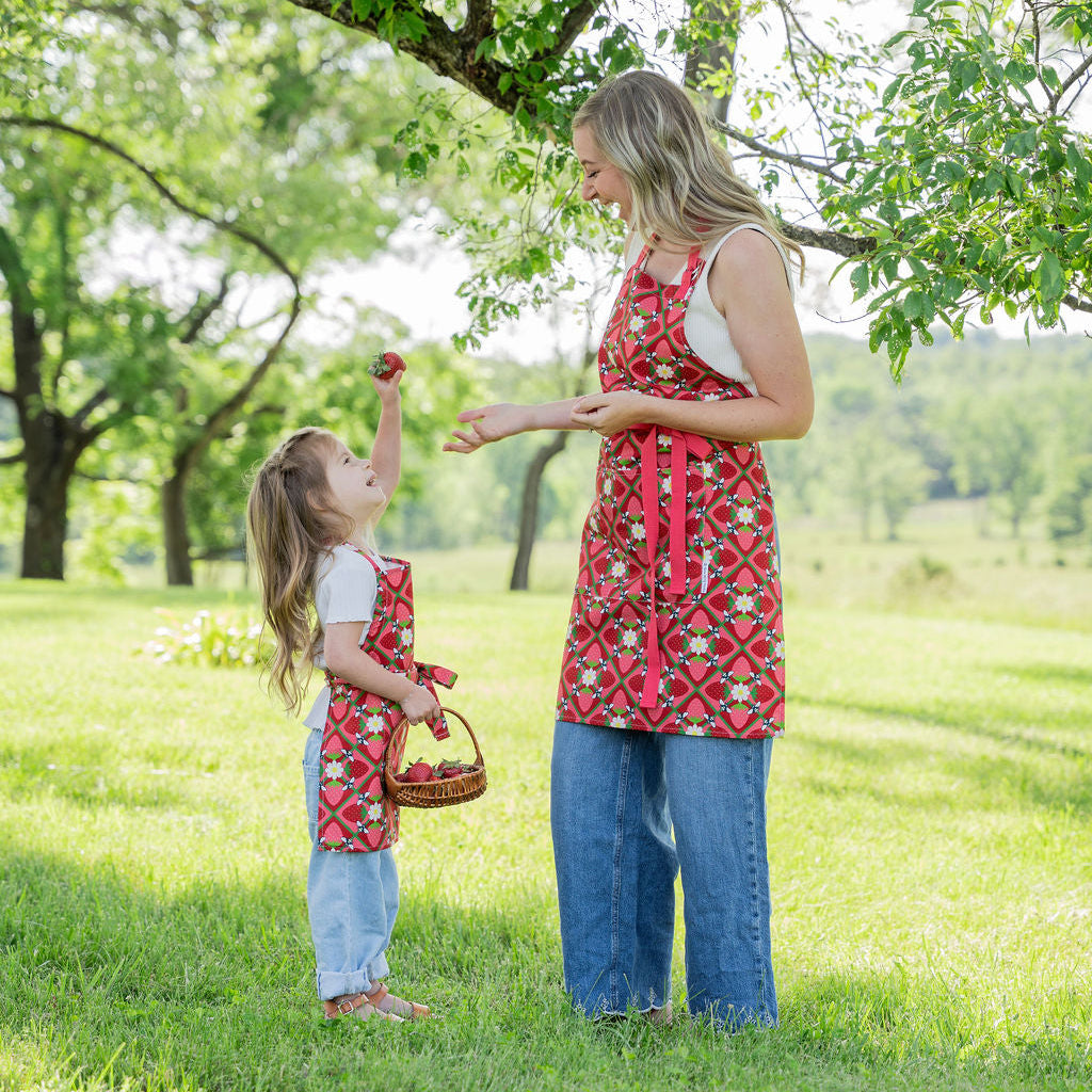 A woman and a young girl wearing matching red patterned aprons stand on a grassy lawn under a tree. The aprons have a red base with a geometric floral pattern in yellow, white, and green.