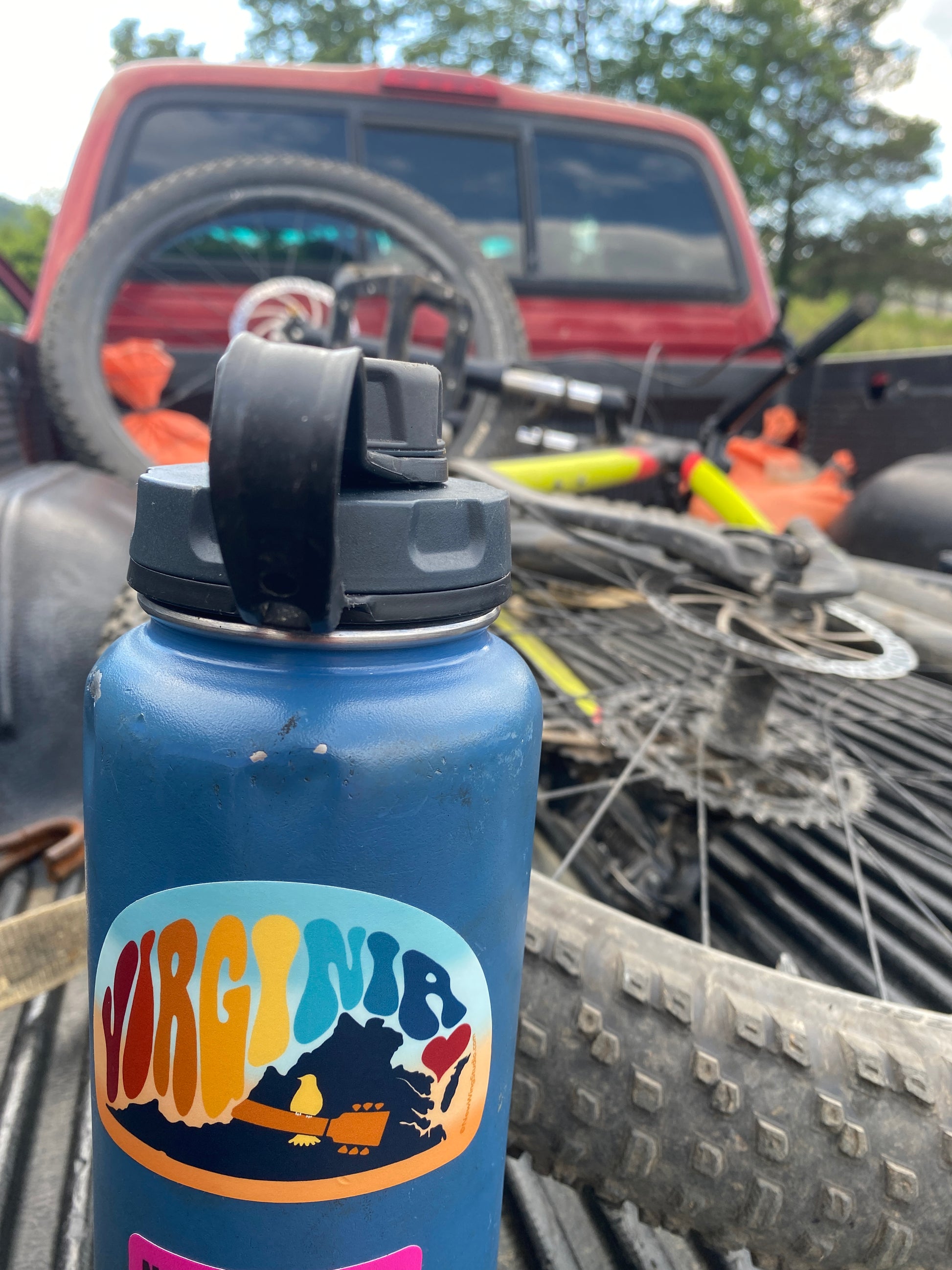 This is a picture of a blue water bottle with a mountain bike in the background, all in the bed of a red pickup truck.  New Wing Studio's Virginia Vibe sticker is displayed on the water bottle.  The sticker has a 60's vibe with large psychedelic letters that say “Virginia.”  There is a Woodstock themed guitar and dove with a heart for “Virginia is for Lovers.”  This retro sticker can be found at newwingstudio.com