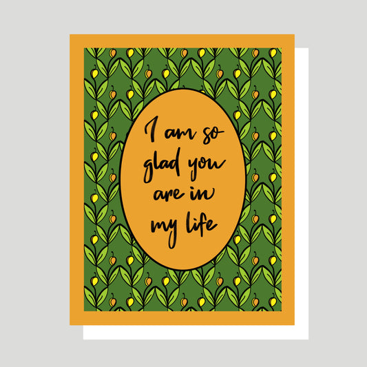 This is a greeting card with the hand written words “I am so glad you are in my life” printed on a tulip inspired pattern with a color scheme that includes a deep orange, a rich green, and a bright yellow.  The words are printed on an oval in the center of the pattern.  The card is beautiful, heart-felt, meaningful, and conveys a message of love, hope, care, and affirmation. The card is printed in Charlottesville Virginia and created by Jennifer Knight, New Wing Studio.